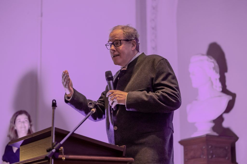 Man speaking at a podium with a microphone, gesturing in a purple-lit room with projected silhouettes behind him