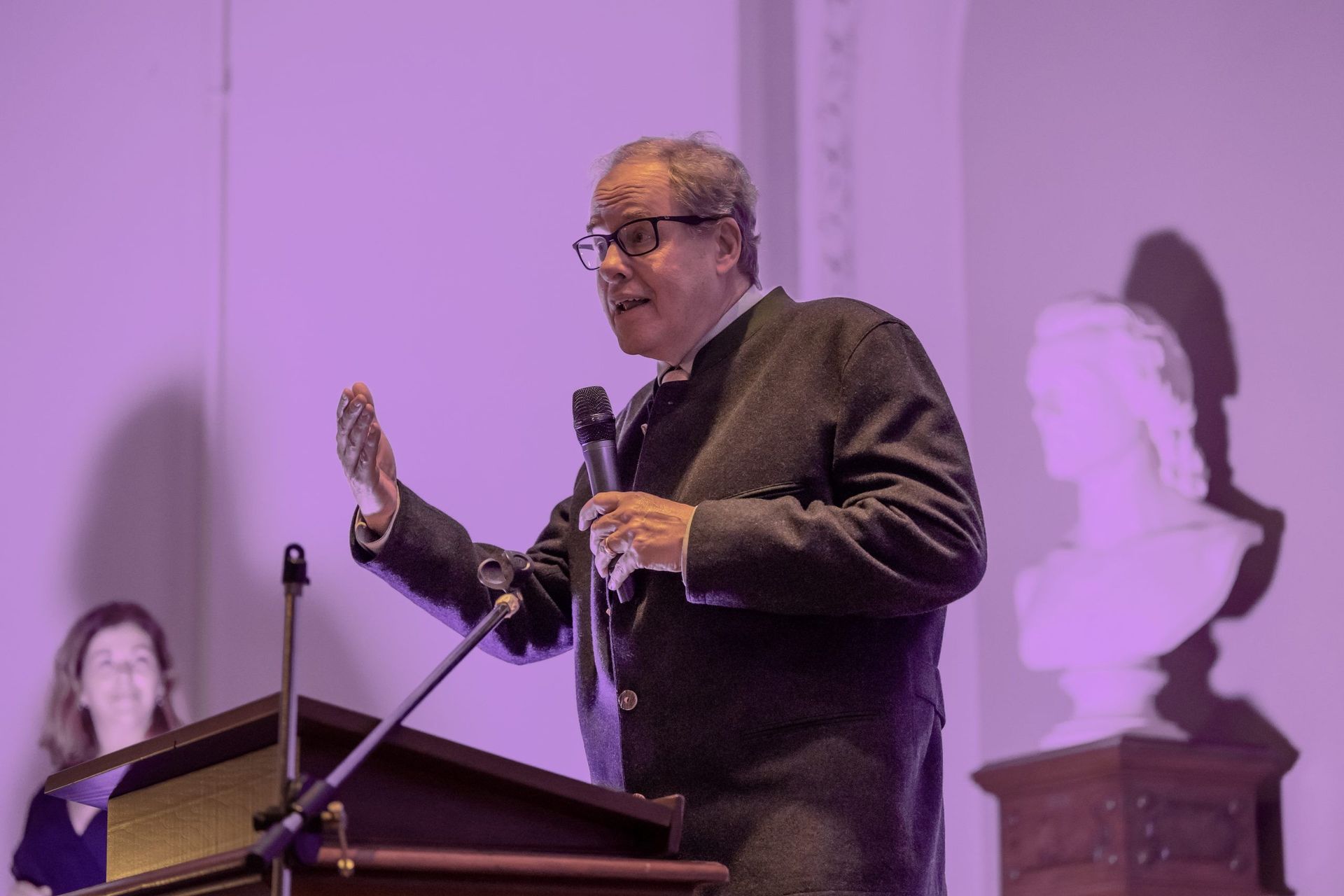 Speaker at a podium in a purple-lit room, gesturing while holding a microphone.