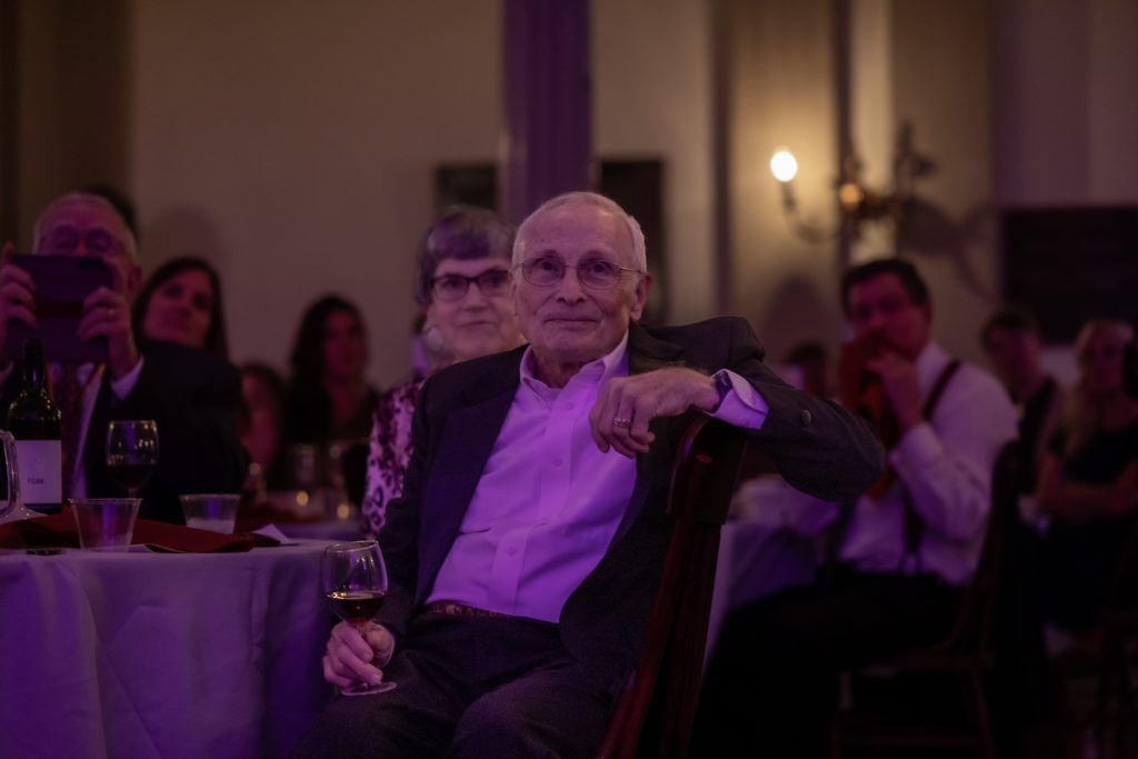 A man in a suit sits at a formal event, holding a glass under purple lighting.