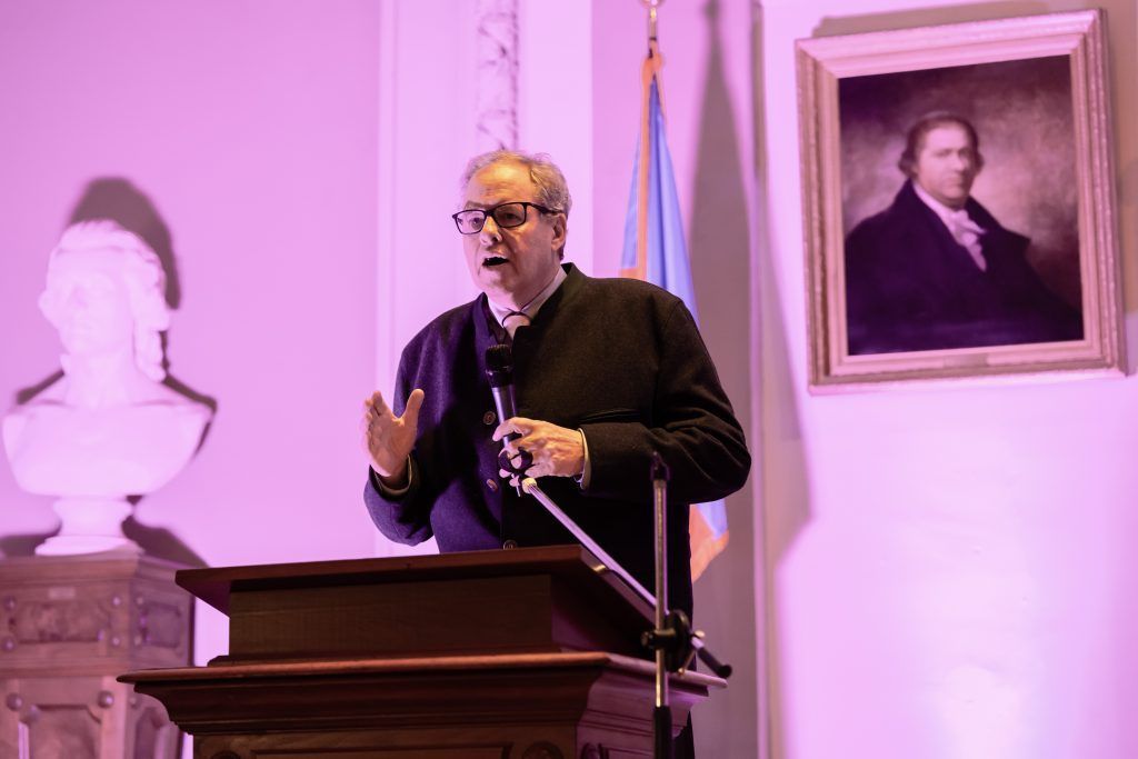 Man speaking at a podium in a purple-lit room, with a bust and framed portrait behind him.