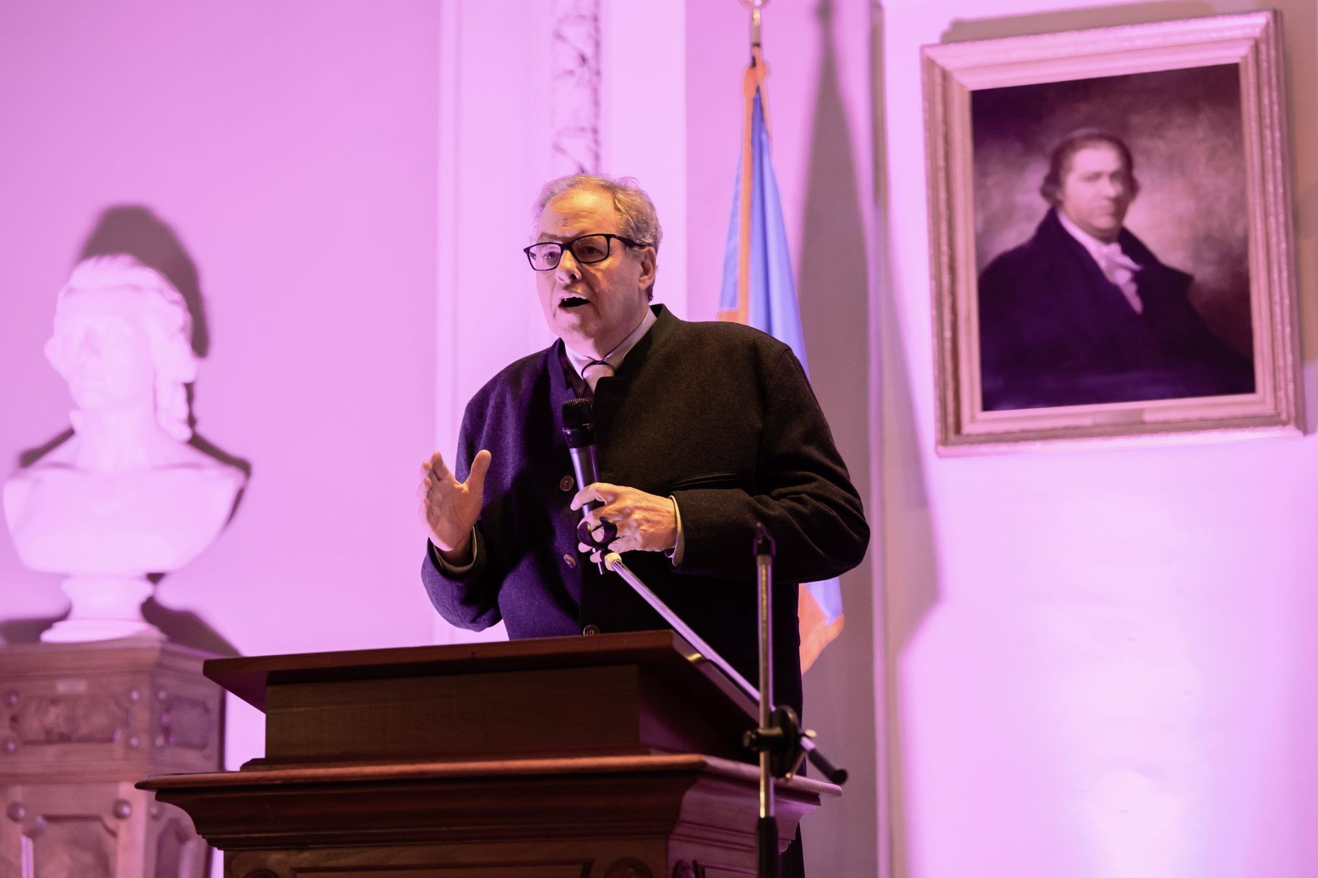 Man speaking at a podium in a purple-lit room with a bust and portrait behind him