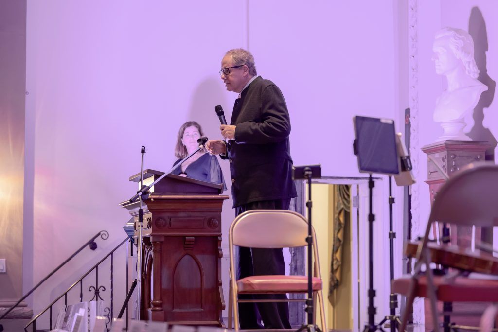 Man speaking at a podium in a church, with a seated person behind him and white statues on the wall