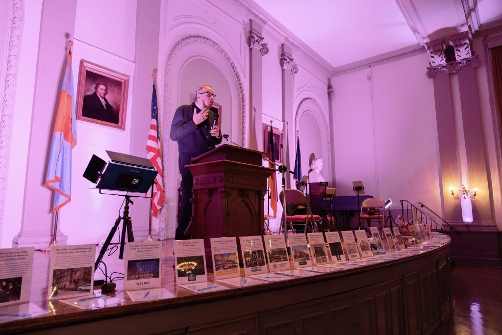 Speaker at a podium in a pink-lit hall with rows of chairs and framed portraits in the background