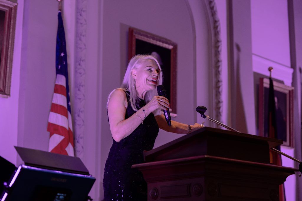 Speaker at podium in a purple-lit hall, gesturing beside a U.S. flag and framed portraits