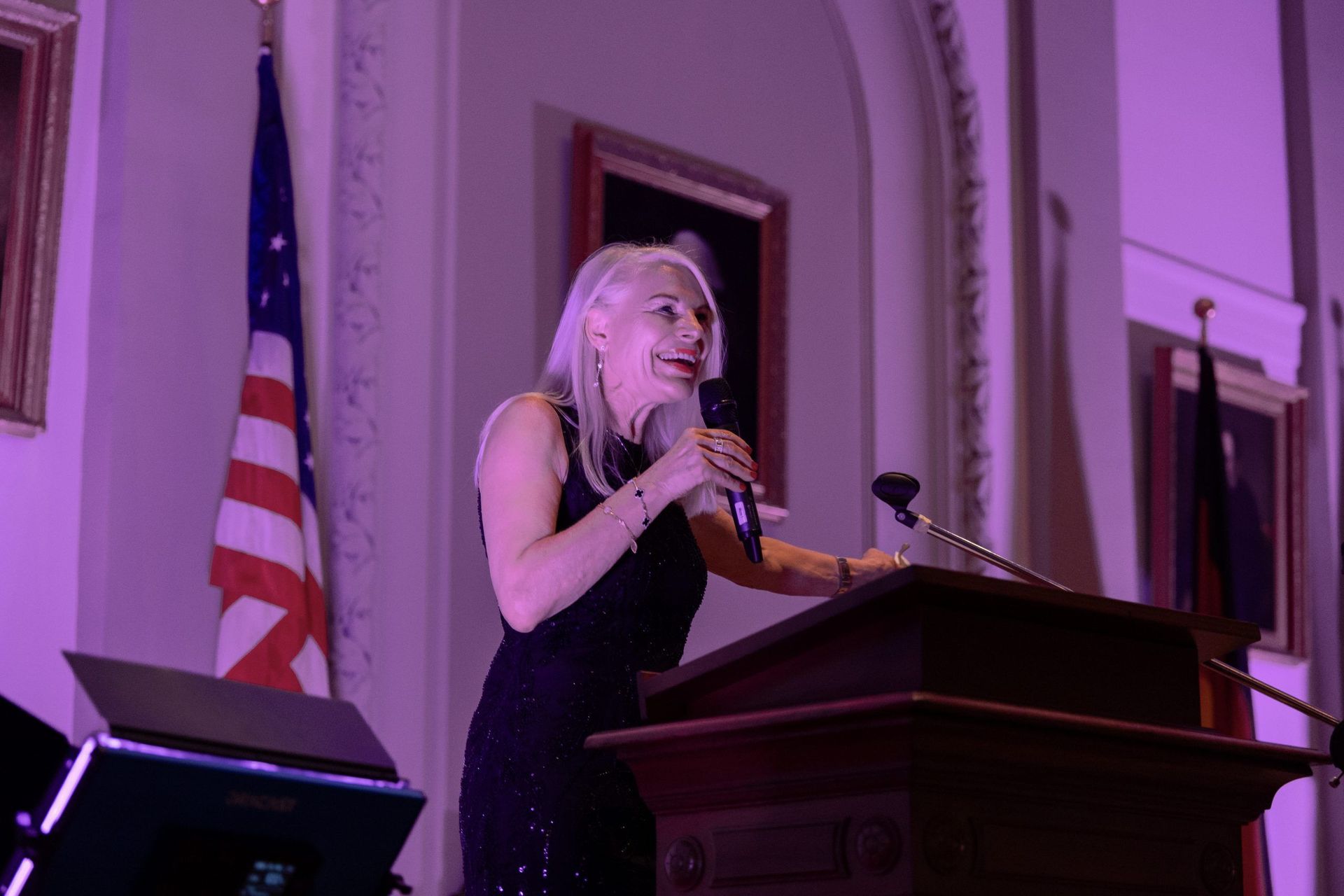 Woman speaking at a podium in a purple-lit hall with a U.S. flag behind her.