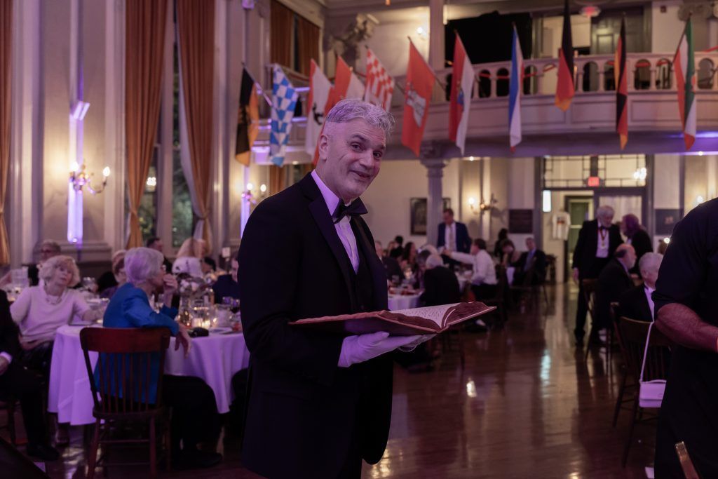 Man holding a tray in a banquet hall with seated guests, flags, and purple lighting