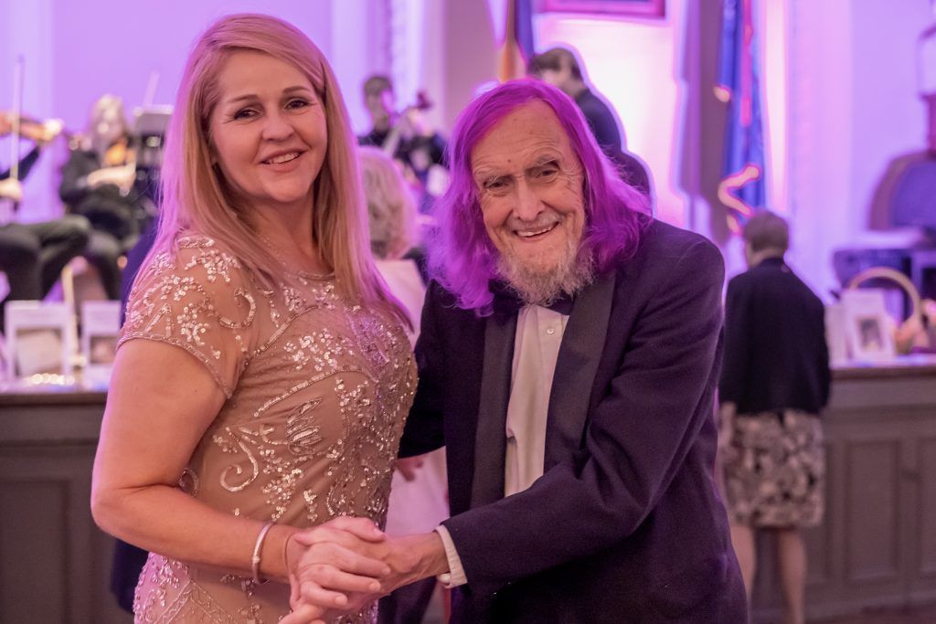 Two people pose smiling and holding hands at a formal event with purple lighting.