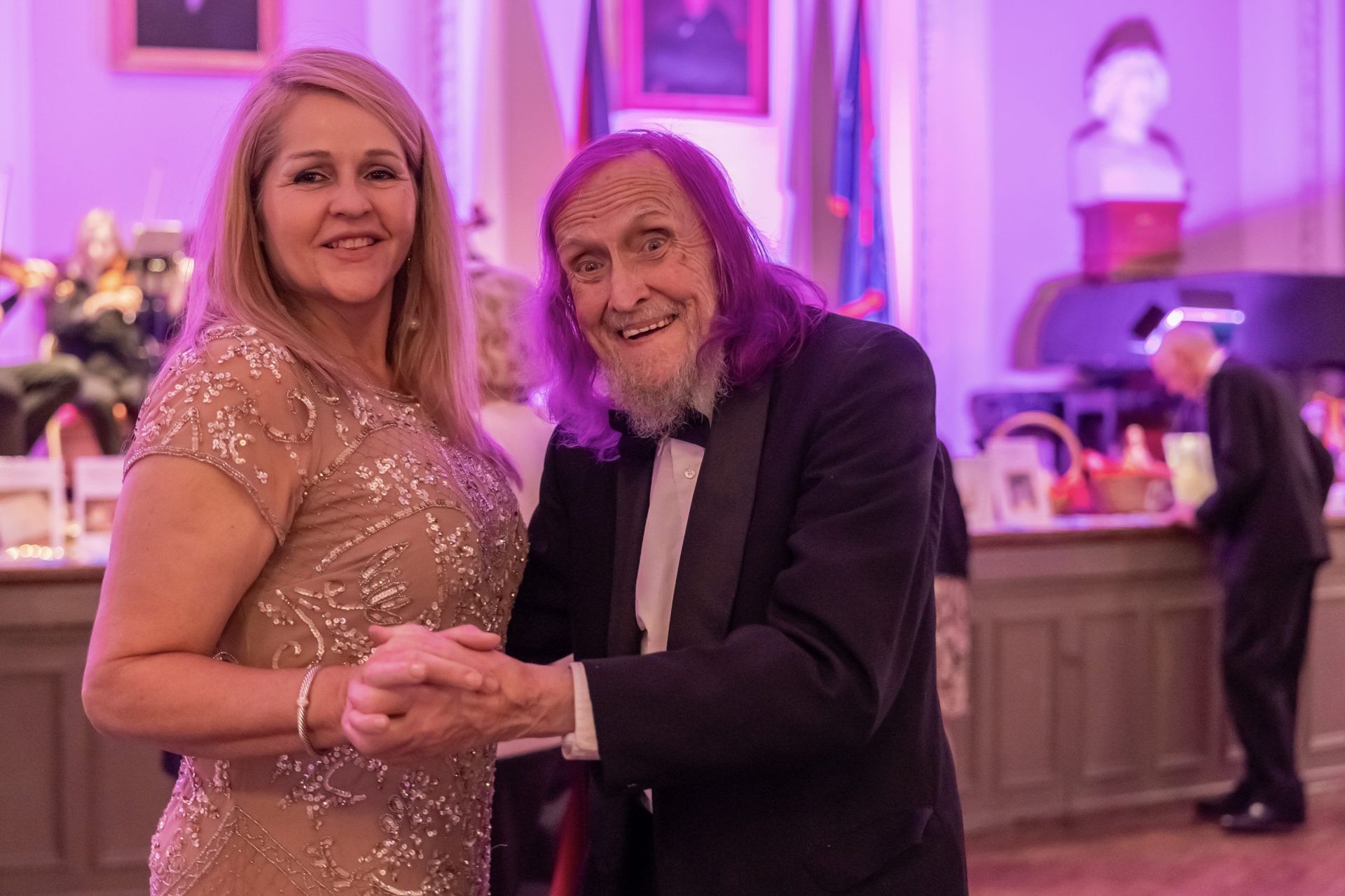 Couple dancing at a formal event, smiling under pink-purple lighting.