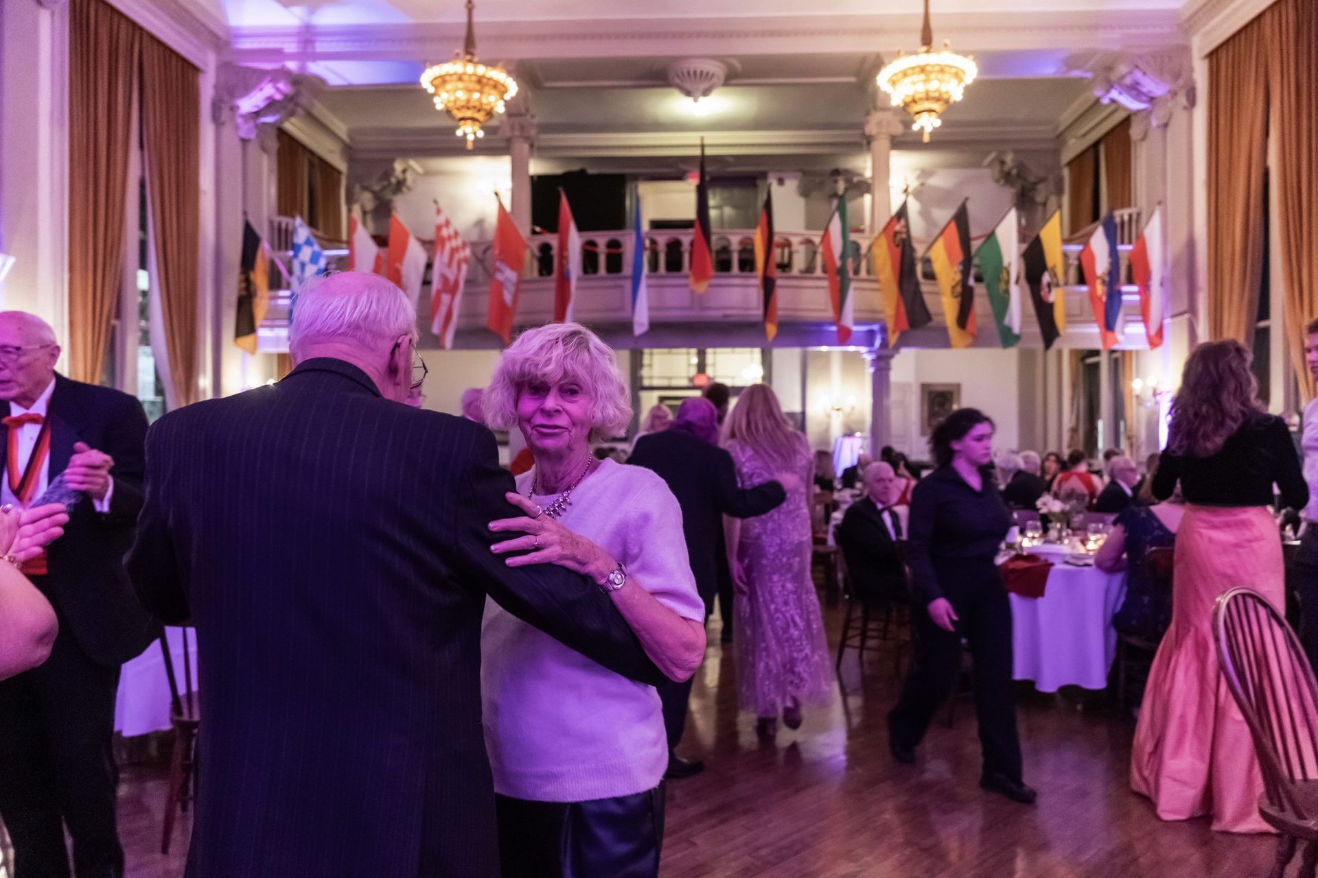 People dancing at a formal event in a chandelier-lit ballroom, with guests mingling at tables.