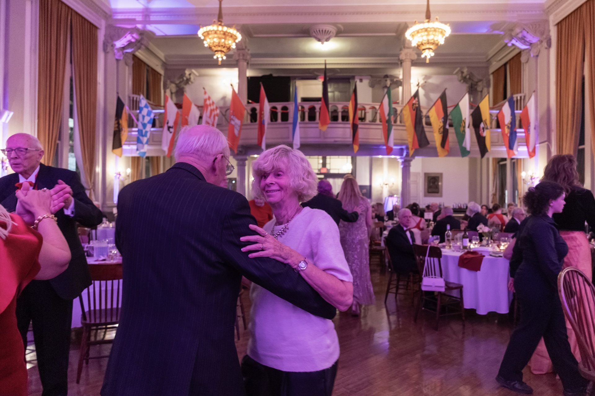 Couple dancing at a formal event in a ballroom with flags and round tables in the background