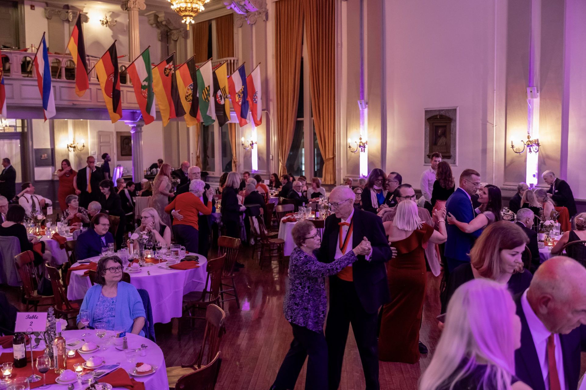 Guests dance and mingle at a warmly lit banquet hall with round tables and hanging flags.