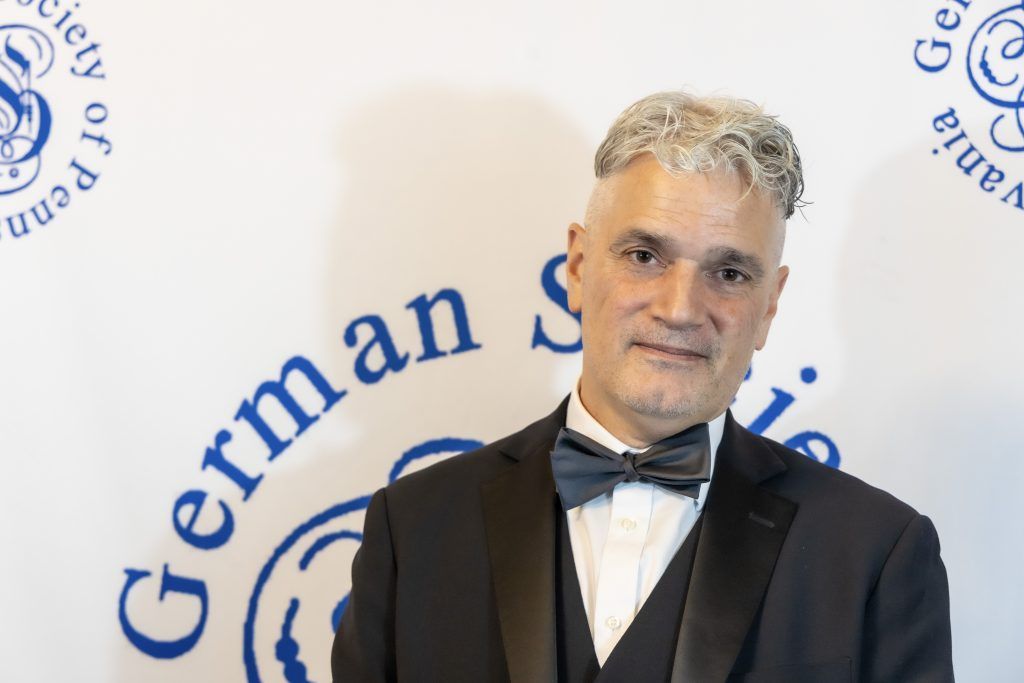 Man in a black tuxedo and bow tie posing before a German Society backdrop