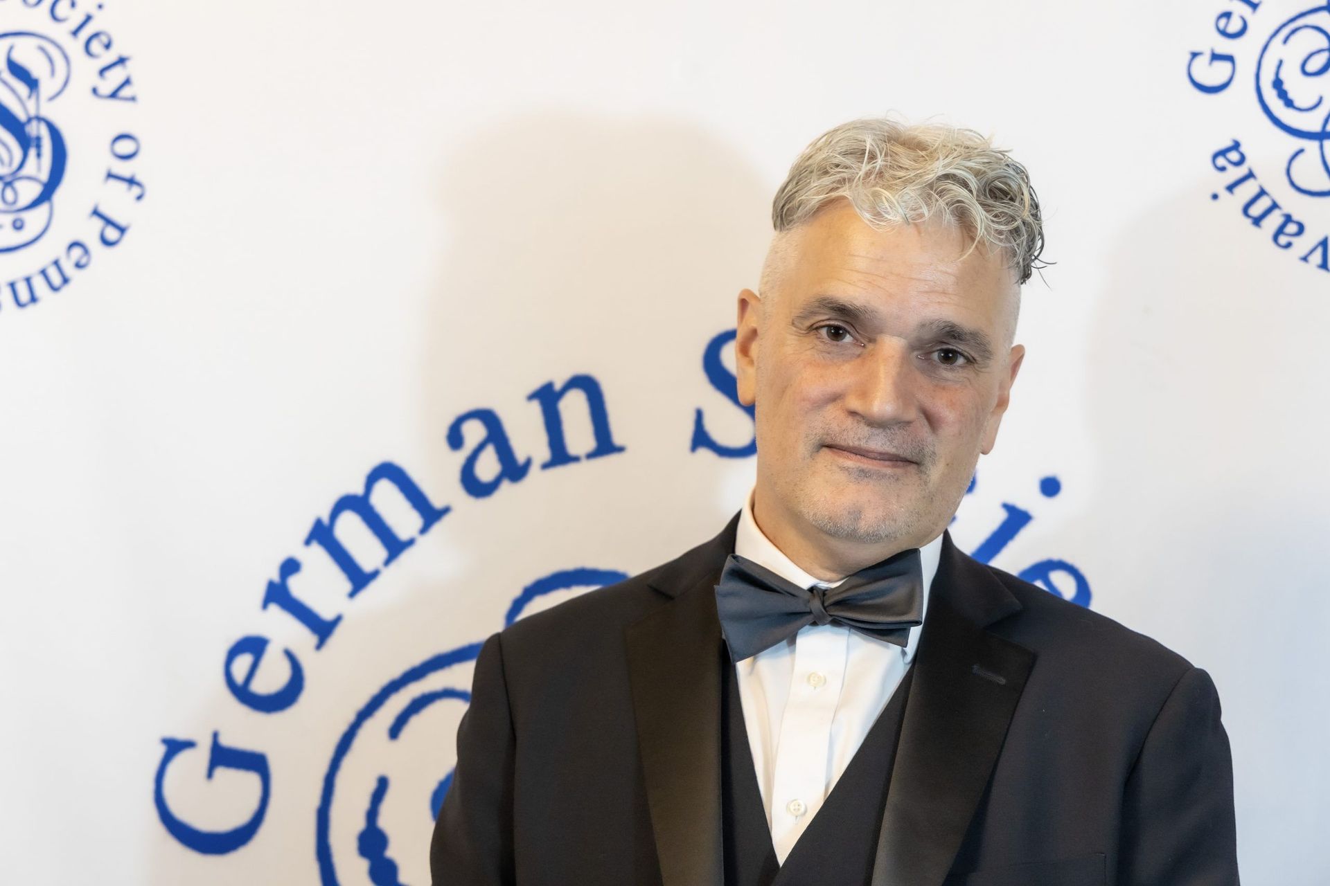 Man in a black tuxedo and bow tie standing before a German Society backdrop