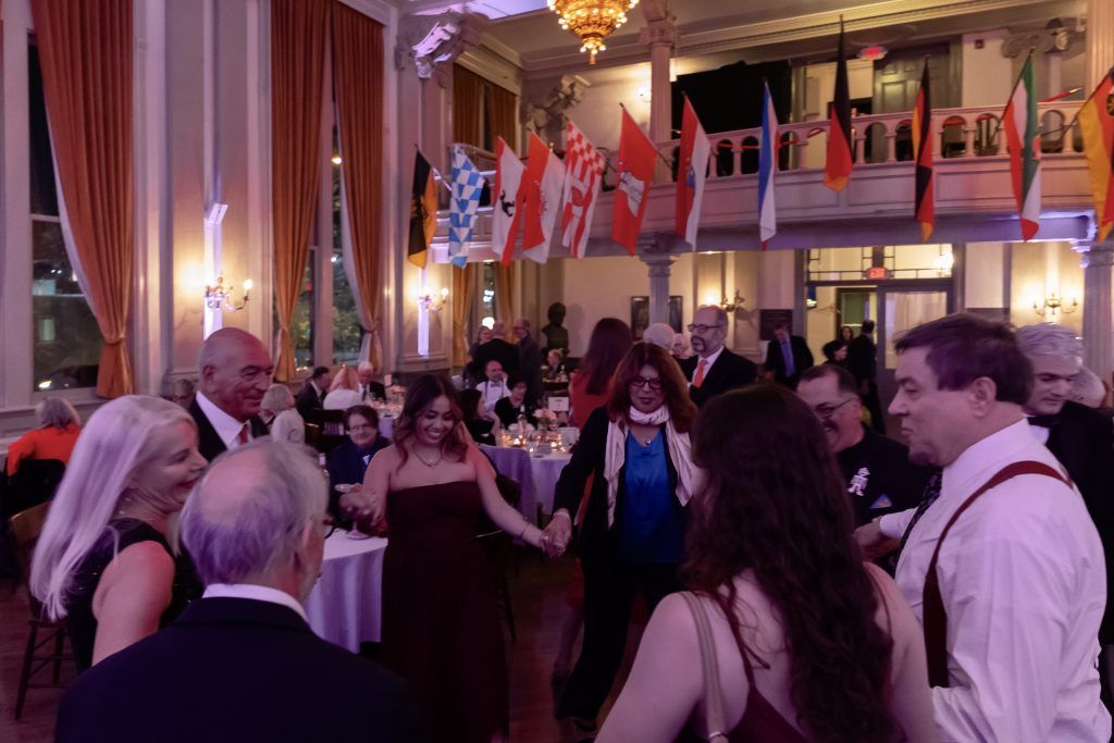 People mingling at a formal indoor event under chandeliers, with international flags overhead.