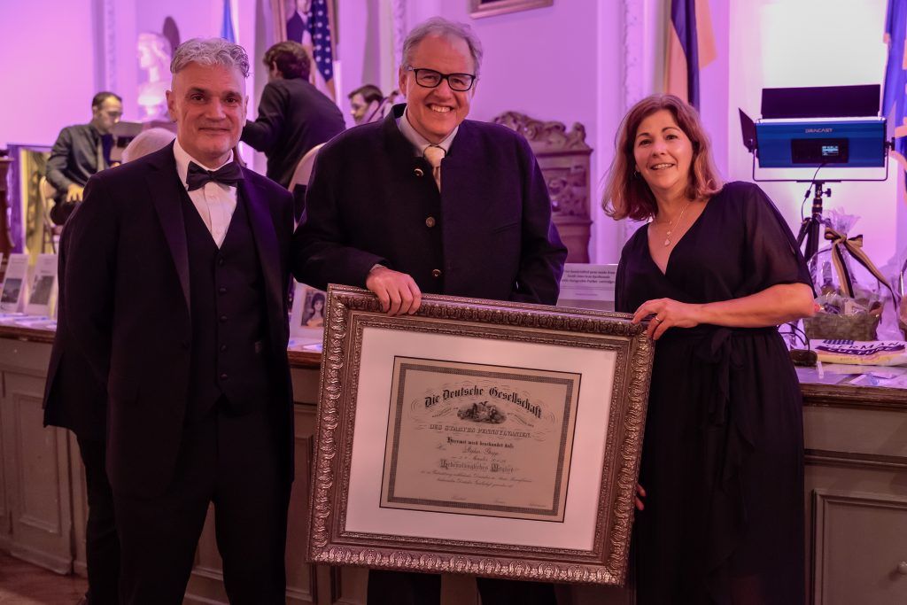 Three people posing with a framed certificate at a purple-lit formal event