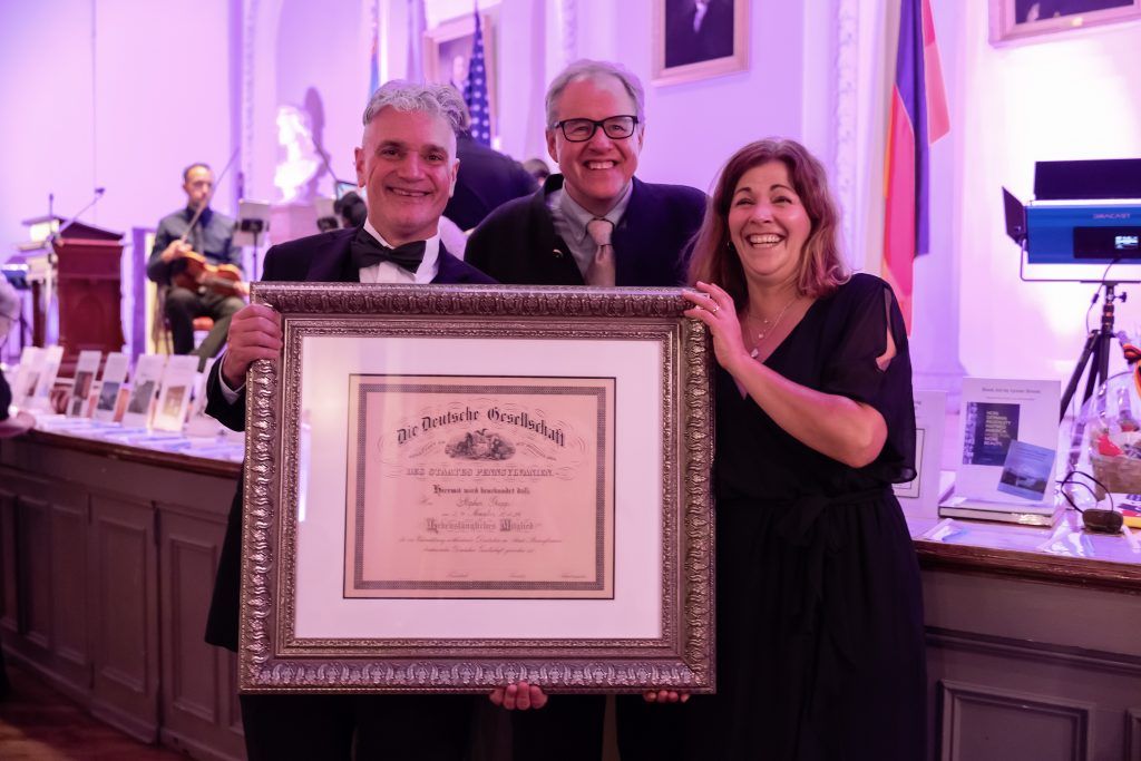 Three people smiling, holding a framed certificate at a banquet table under purple lighting.