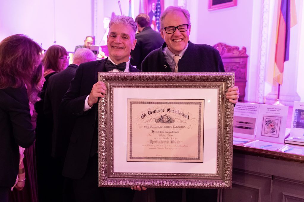 Two men holding a framed certificate at a formal indoor event with pink-purple lighting