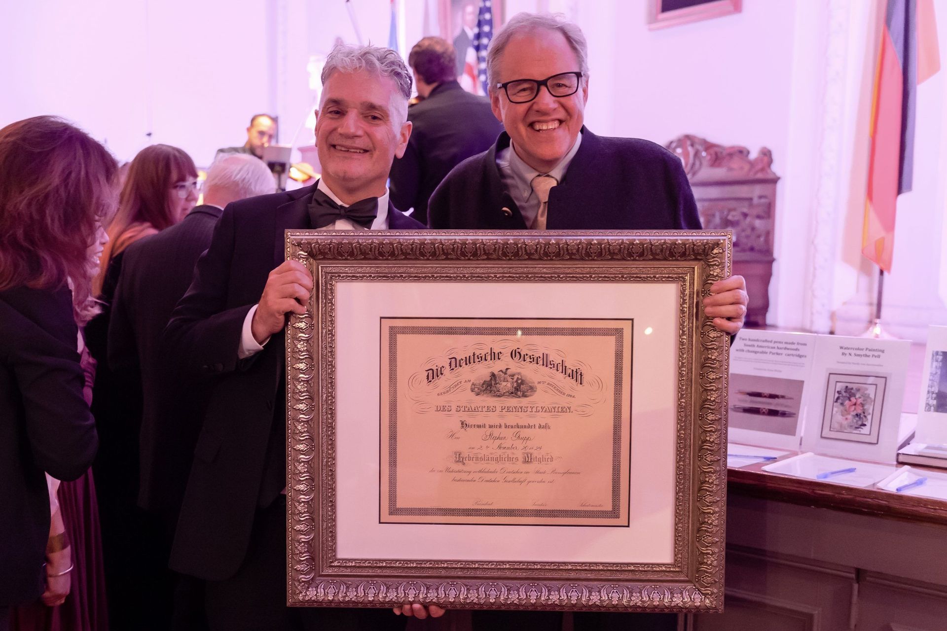 Two people holding a framed certificate at an indoor event, smiling beside a display table.