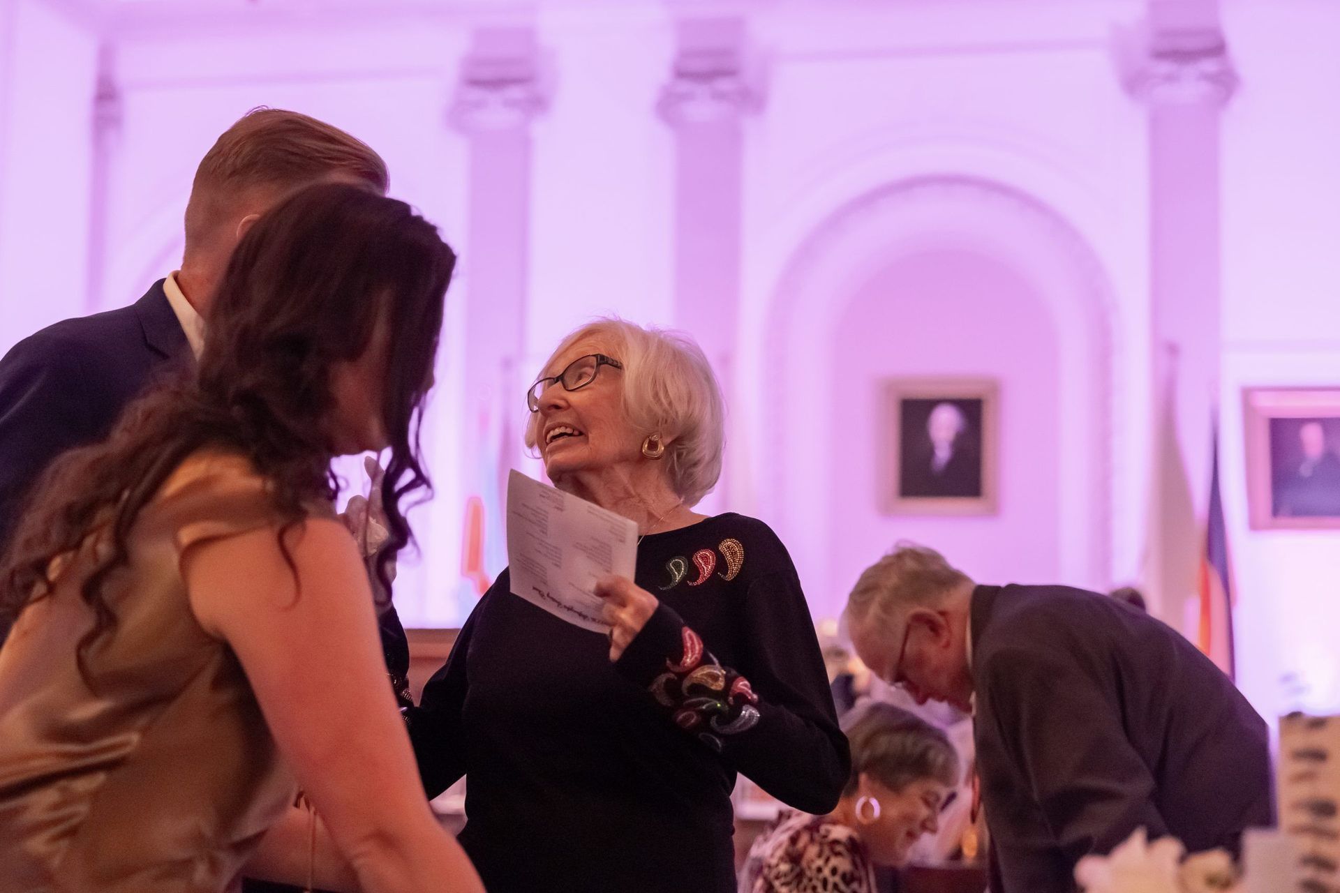 Guests mingling at a purple-lit indoor event, with one woman holding papers and speaking to others.