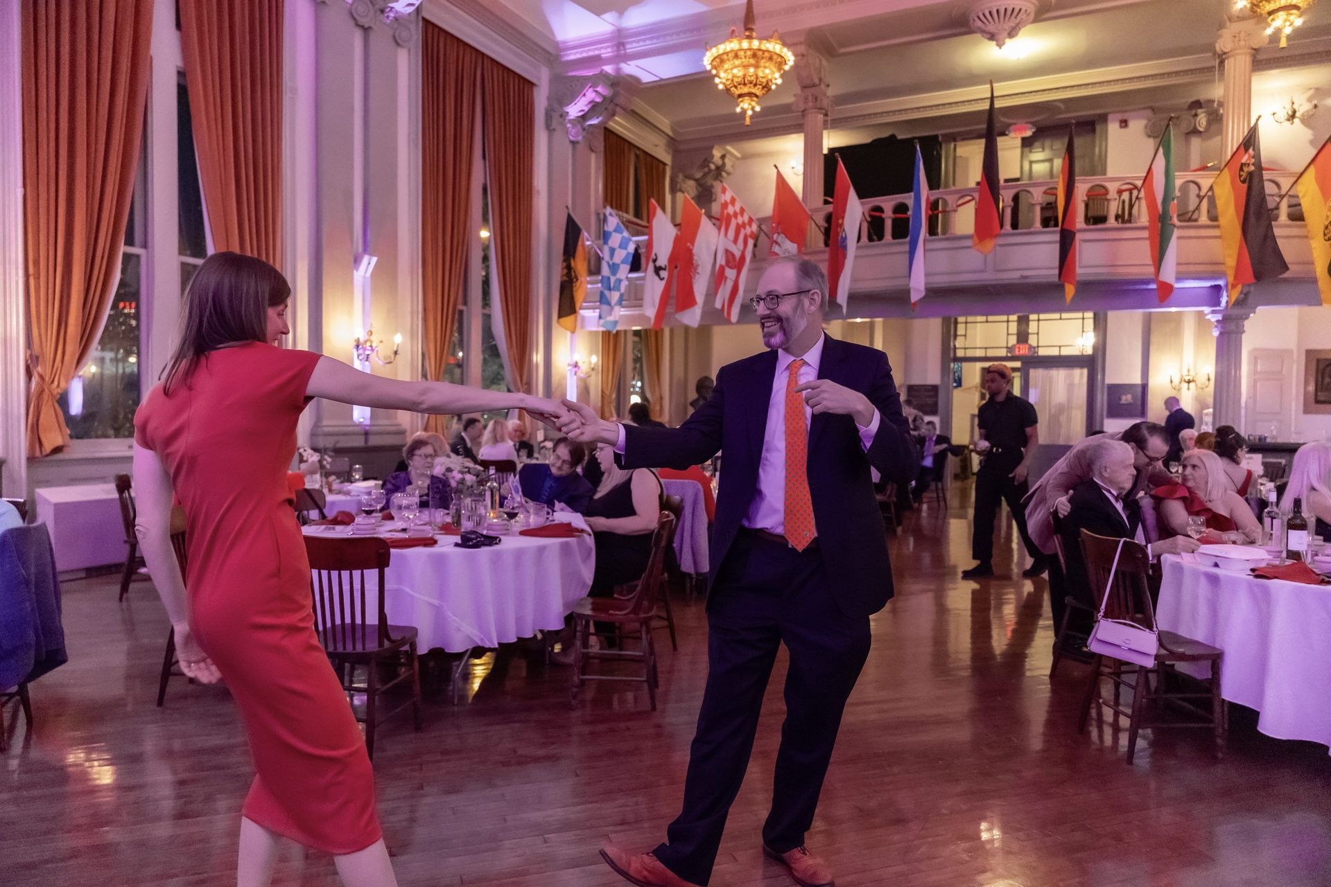 Two people dancing in a banquet hall with round tables, chandeliers, and flags in the background