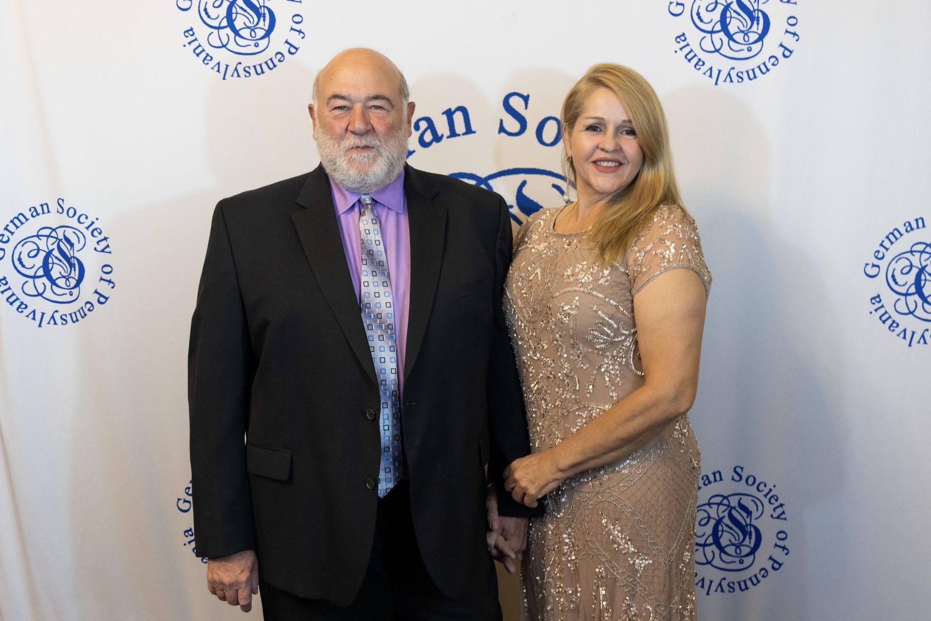 Two people posing on a step-and-repeat backdrop, man in black suit and woman in gold dress