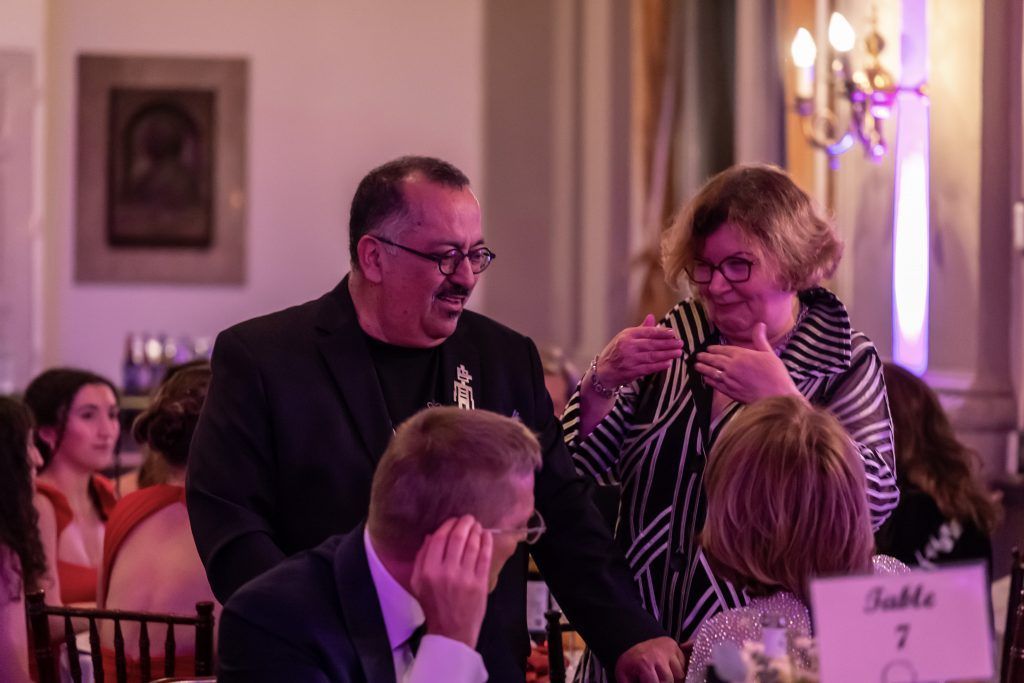 Two people speaking into microphones at a formal indoor event with seated guests nearby.