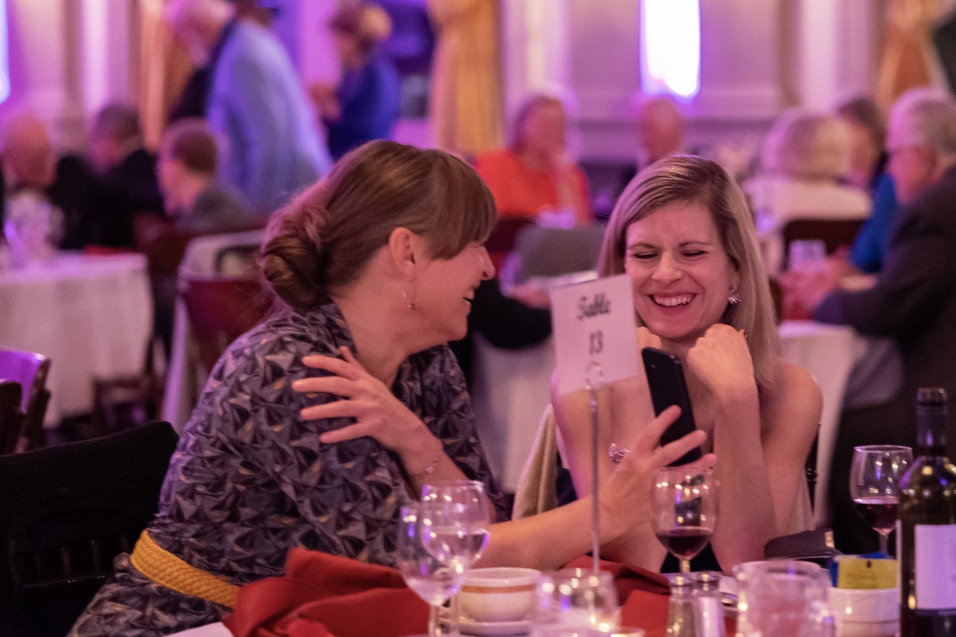Two women laughing at a table during a purple-lit banquet, one holding up a phone