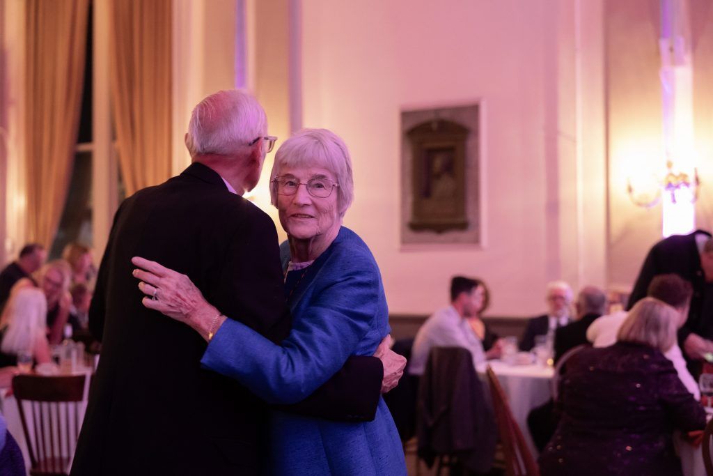 Two people slow dance at a formal event in a softly lit banquet hall.