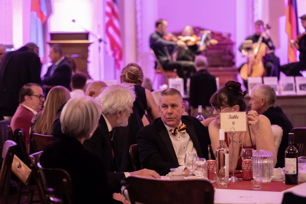 Guests seated at round tables in a banquet hall, with musicians performing onstage in the background.