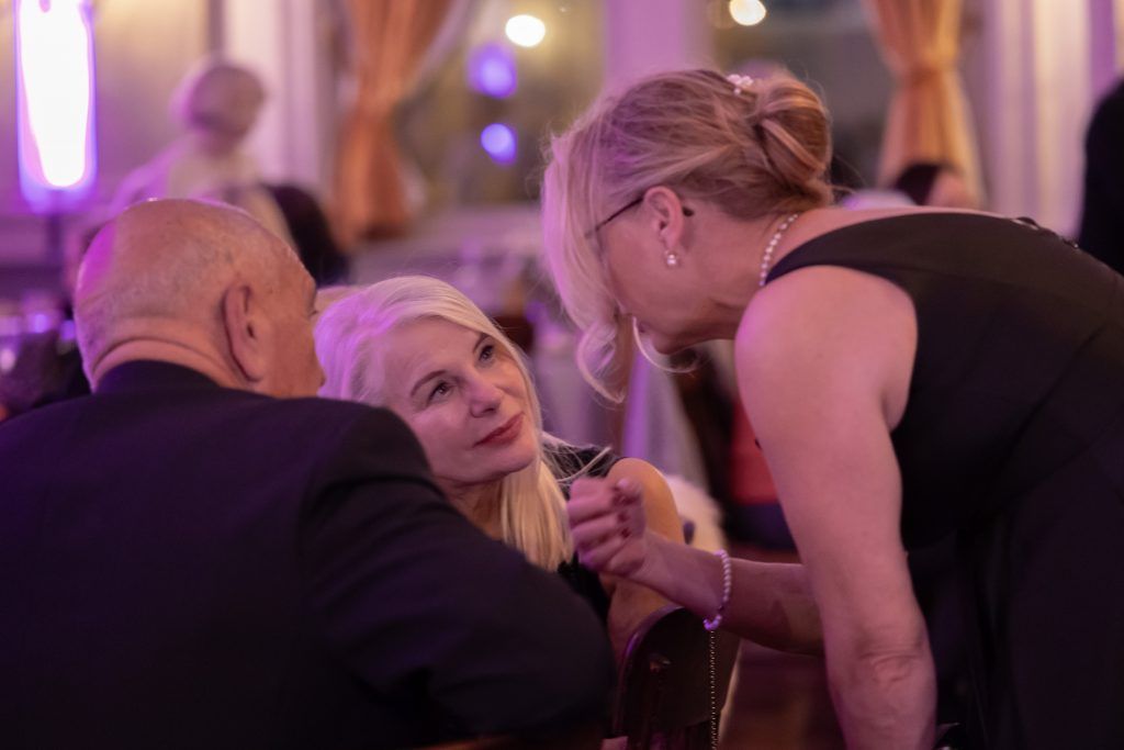 Three people converse at a dimly lit party, one leaning in with a drink in hand.