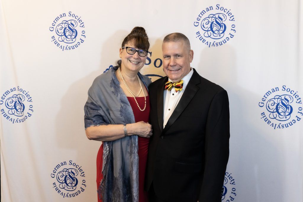 Two people smiling at a formal event, standing before a white step-and-repeat backdrop with blue logos.