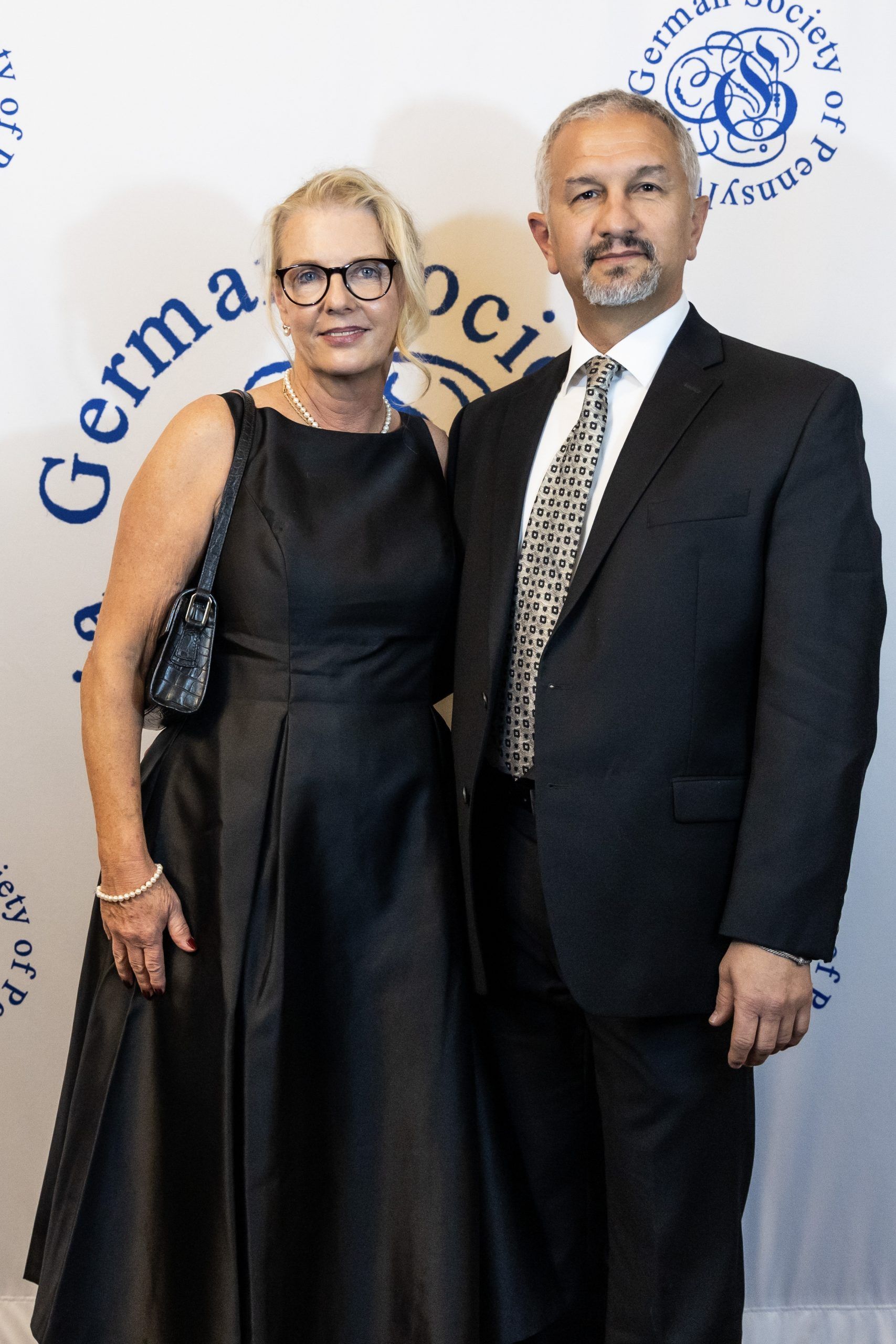 Two people posing at a formal event, wearing black evening attire, in front of a branded backdrop.