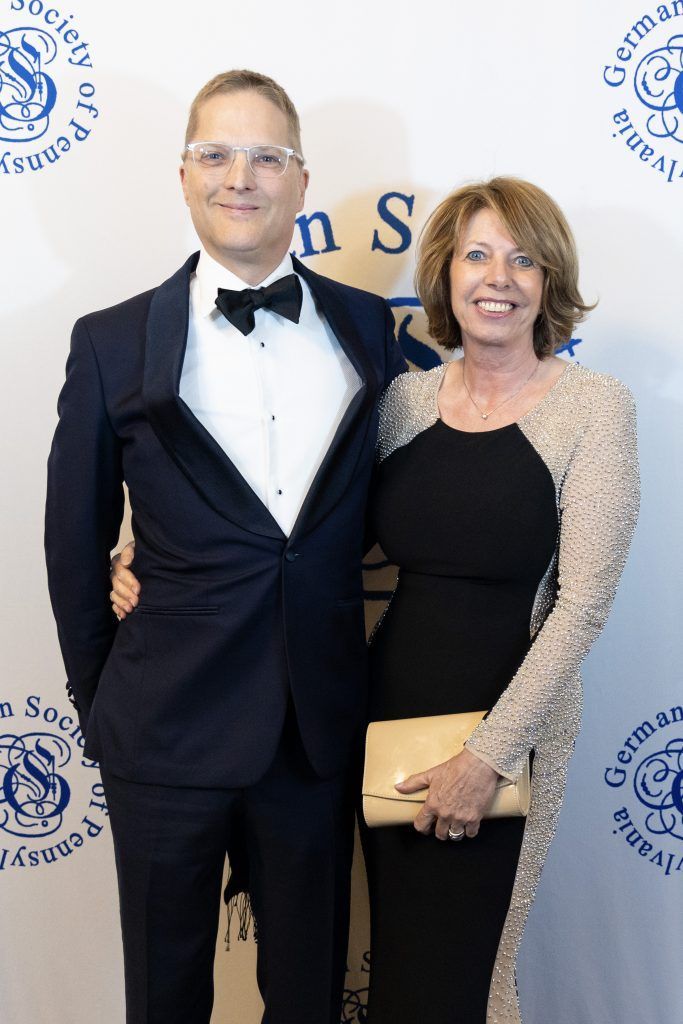 Two people posing at a formal event backdrop, wearing tuxedo and black dress with gold sleeves.