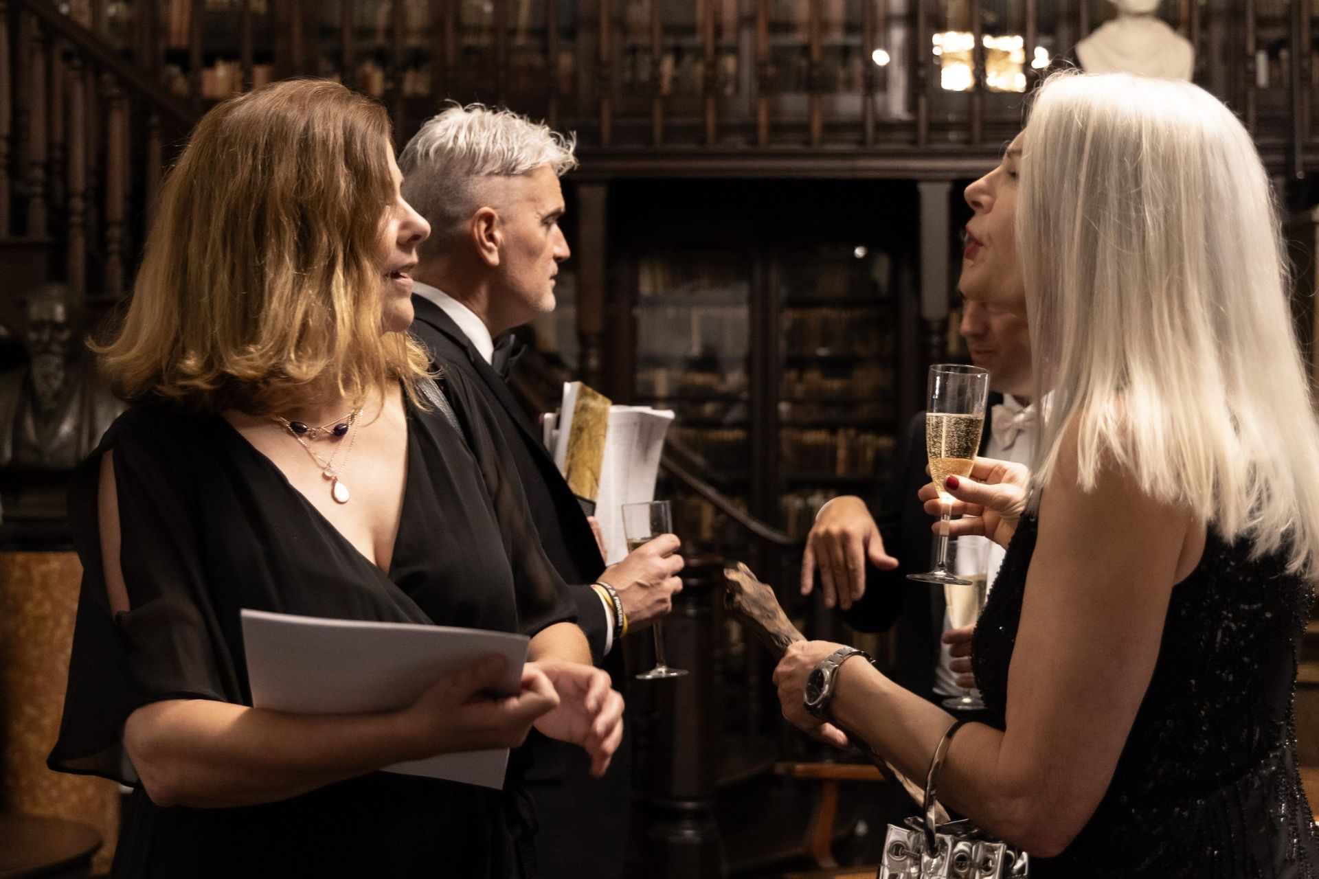 Three people in formal wear chat with drinks in a dimly lit library or study.