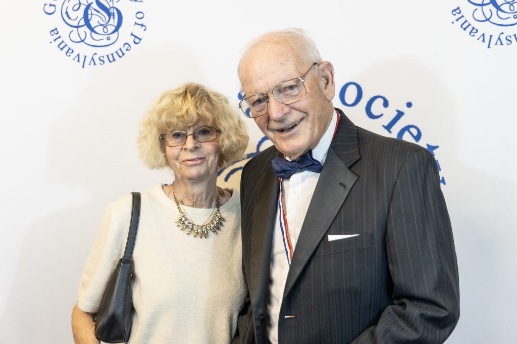 Two smiling people posing together at a formal event, wearing suits and dresses against a branded backdrop.