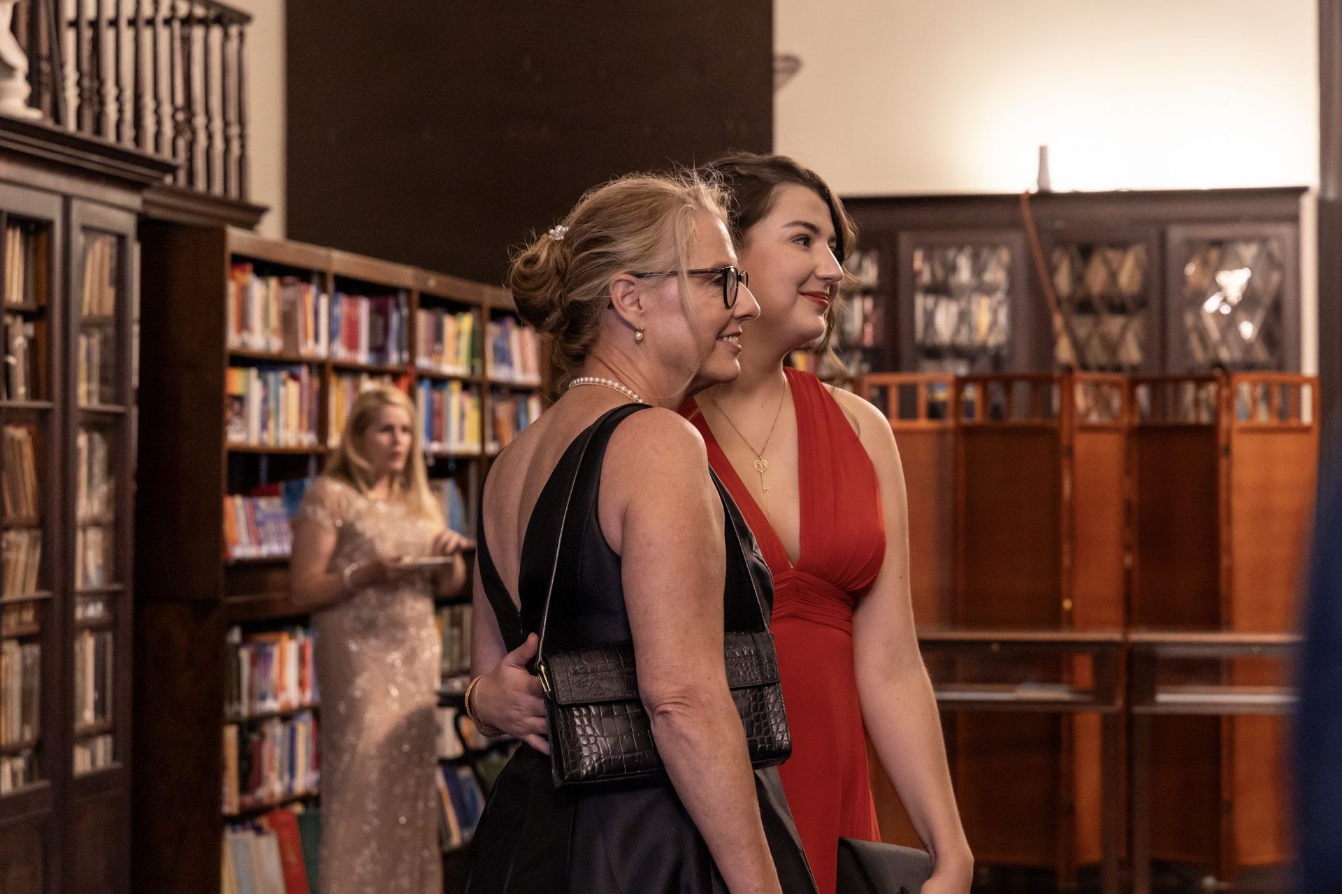 Two women in a library, one in a red dress and one in black, standing and smiling near bookshelves