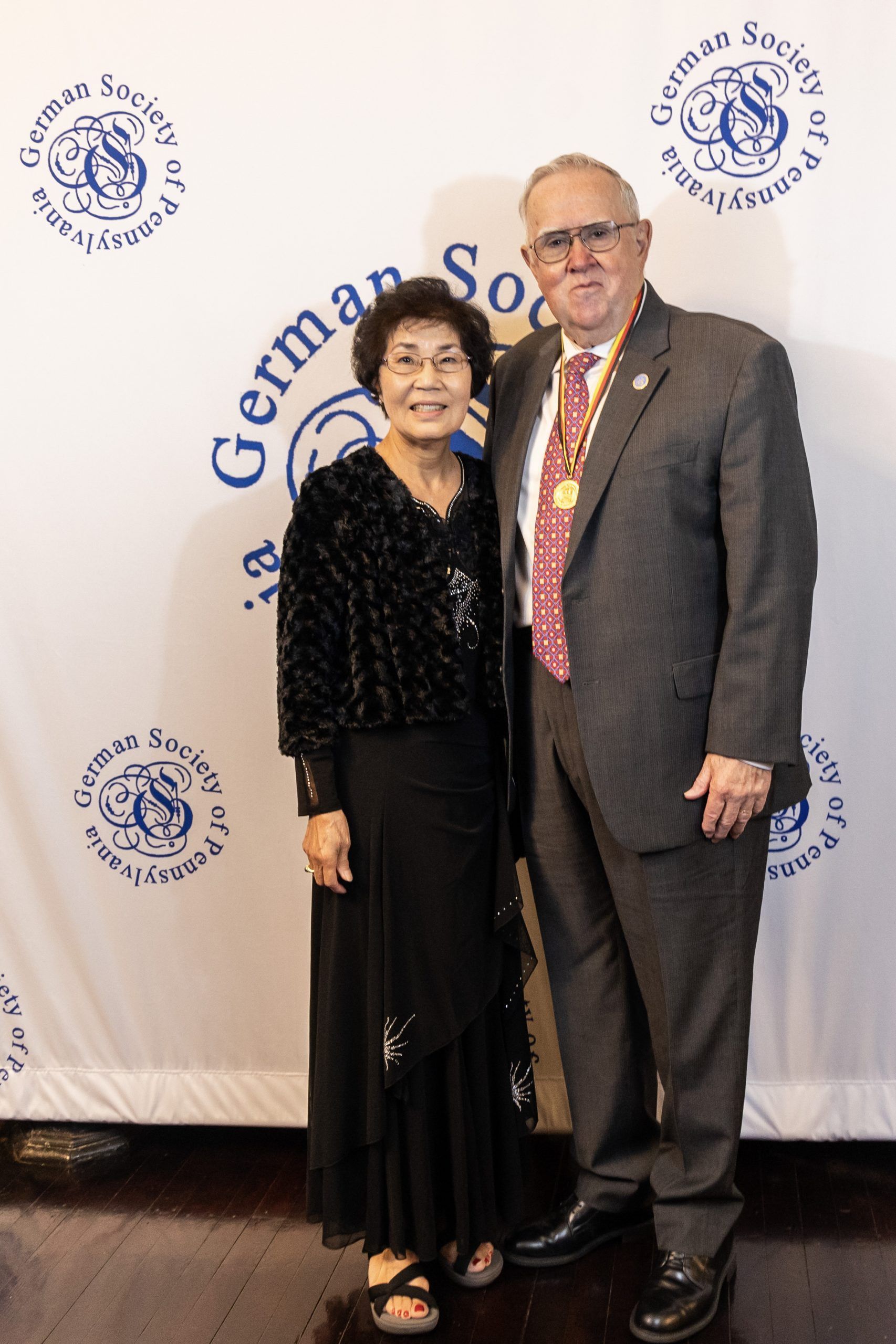 Two people posing at a formal event backdrop, dressed in dark evening attire and smiling.