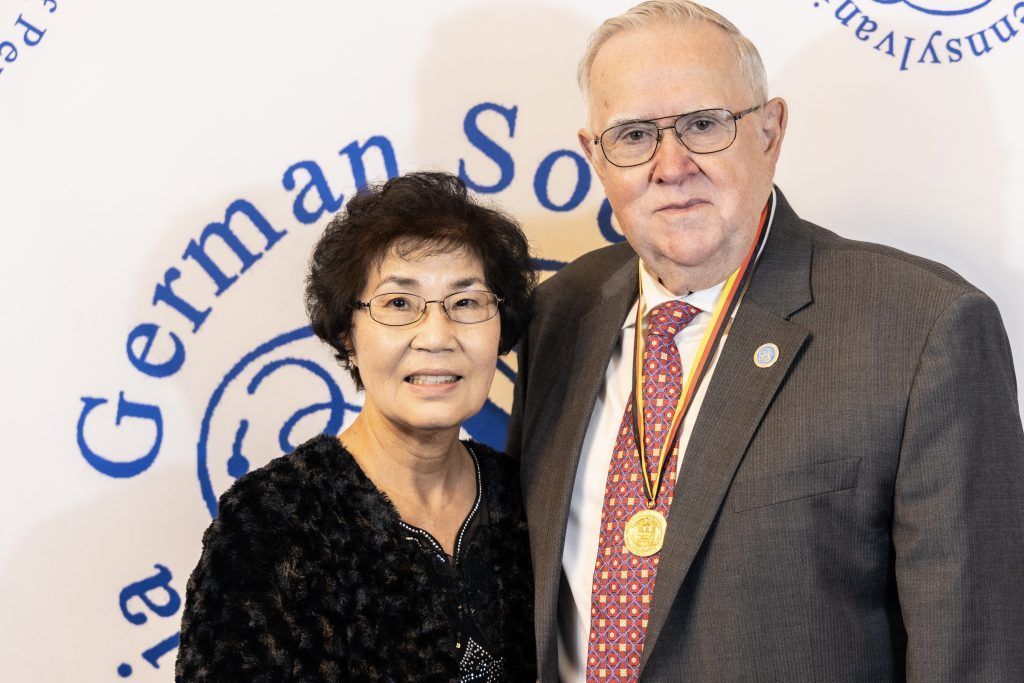 Two people posing at a German Society event backdrop, one wearing a medal.