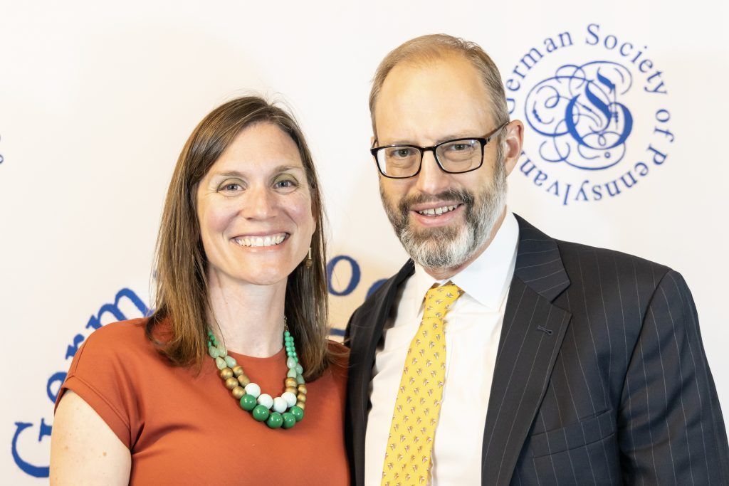 Two smiling people posing in front of a Massachusetts organization backdrop.
