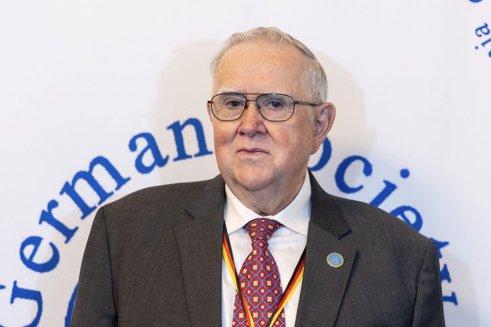 Man in a dark suit and red tie speaking at a podium with German Society logo backdrop