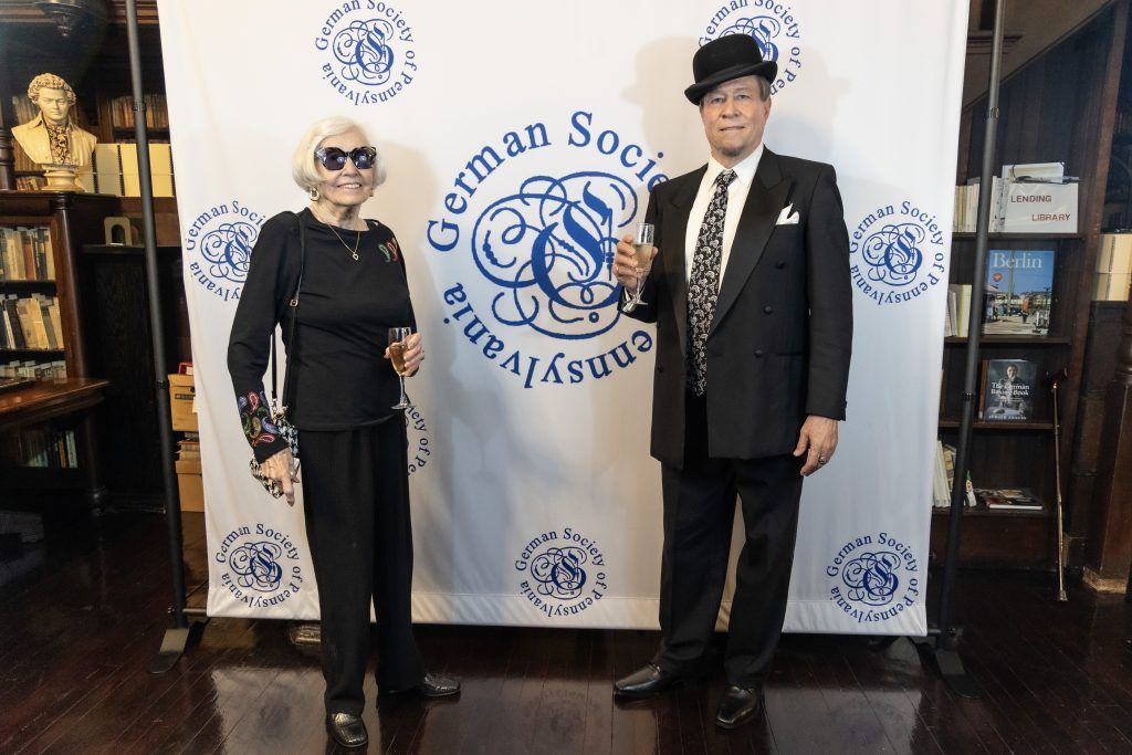 Two people standing indoors in formal black outfits in front of a logo backdrop.