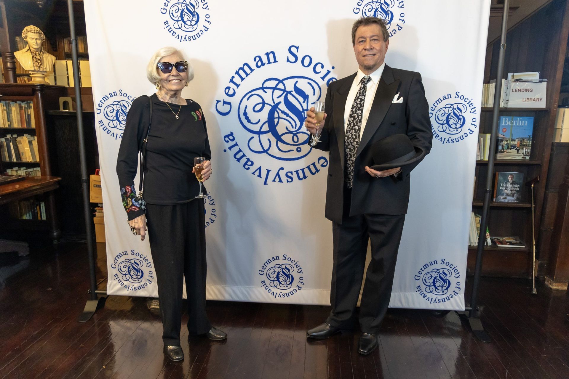 Two people pose in formal wear beside a Grand Society banner in a library setting.