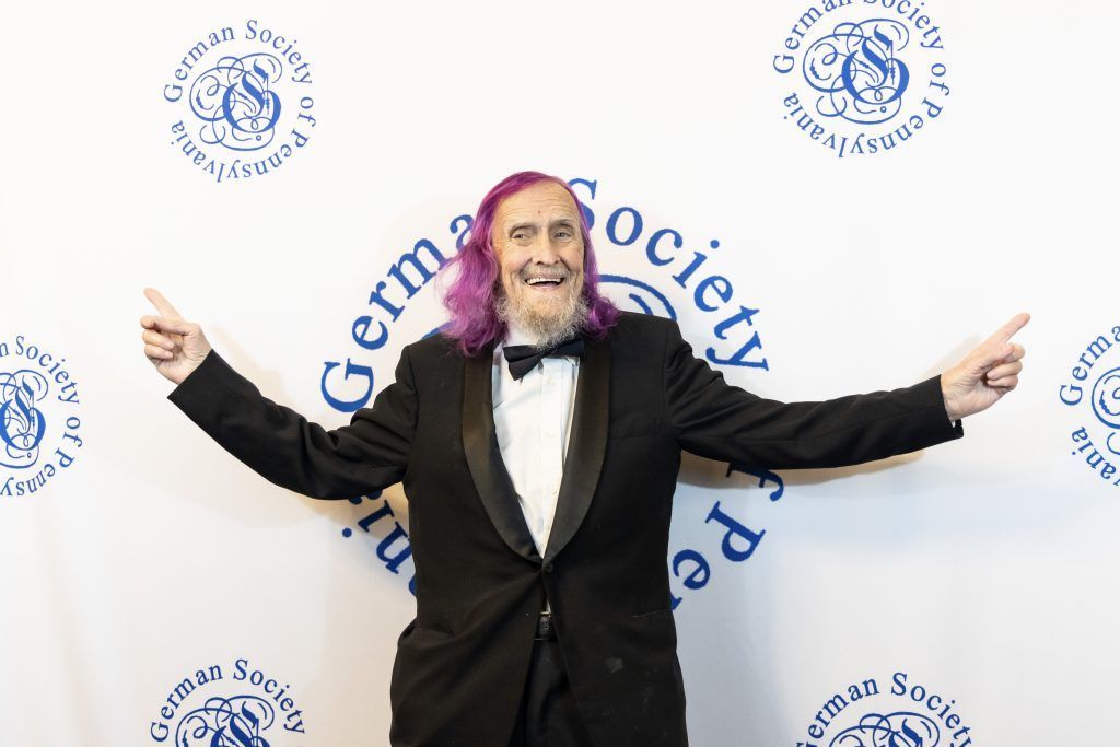 Man in a black tuxedo with arms outstretched at a German Society event backdrop