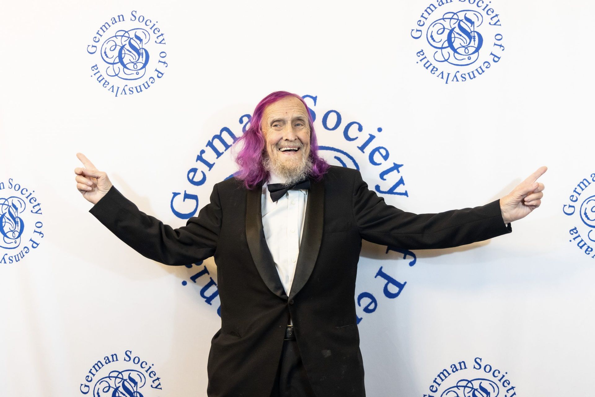 Man in a black tuxedo with outstretched arms posing before a German Society backdrop