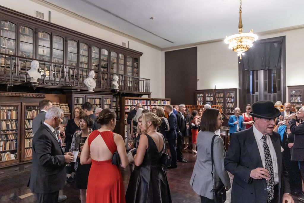 Guests mingle at a formal event in a grand room with glass cabinets and chandeliers.