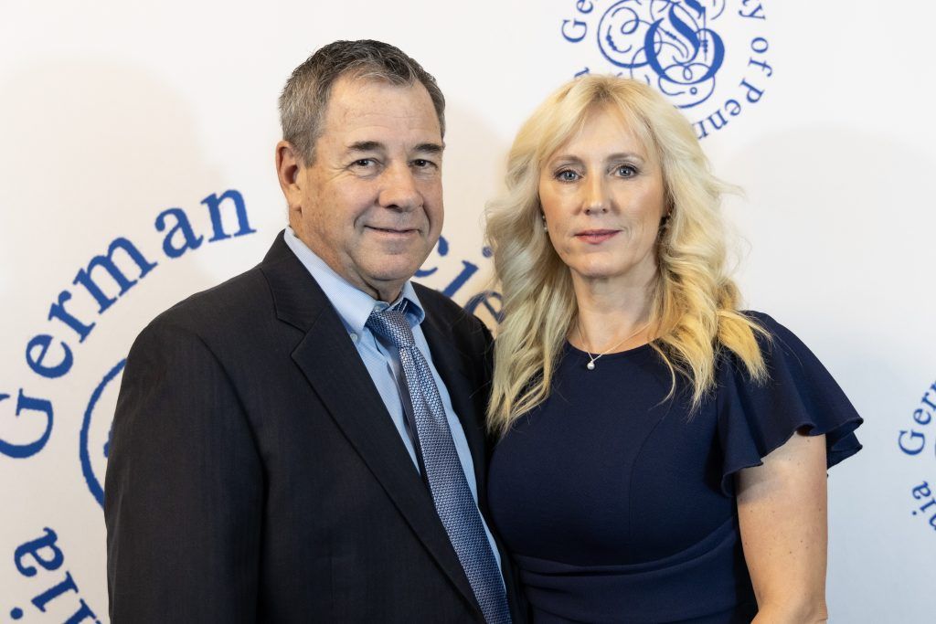 Two people posing in front of a white backdrop with blue German text