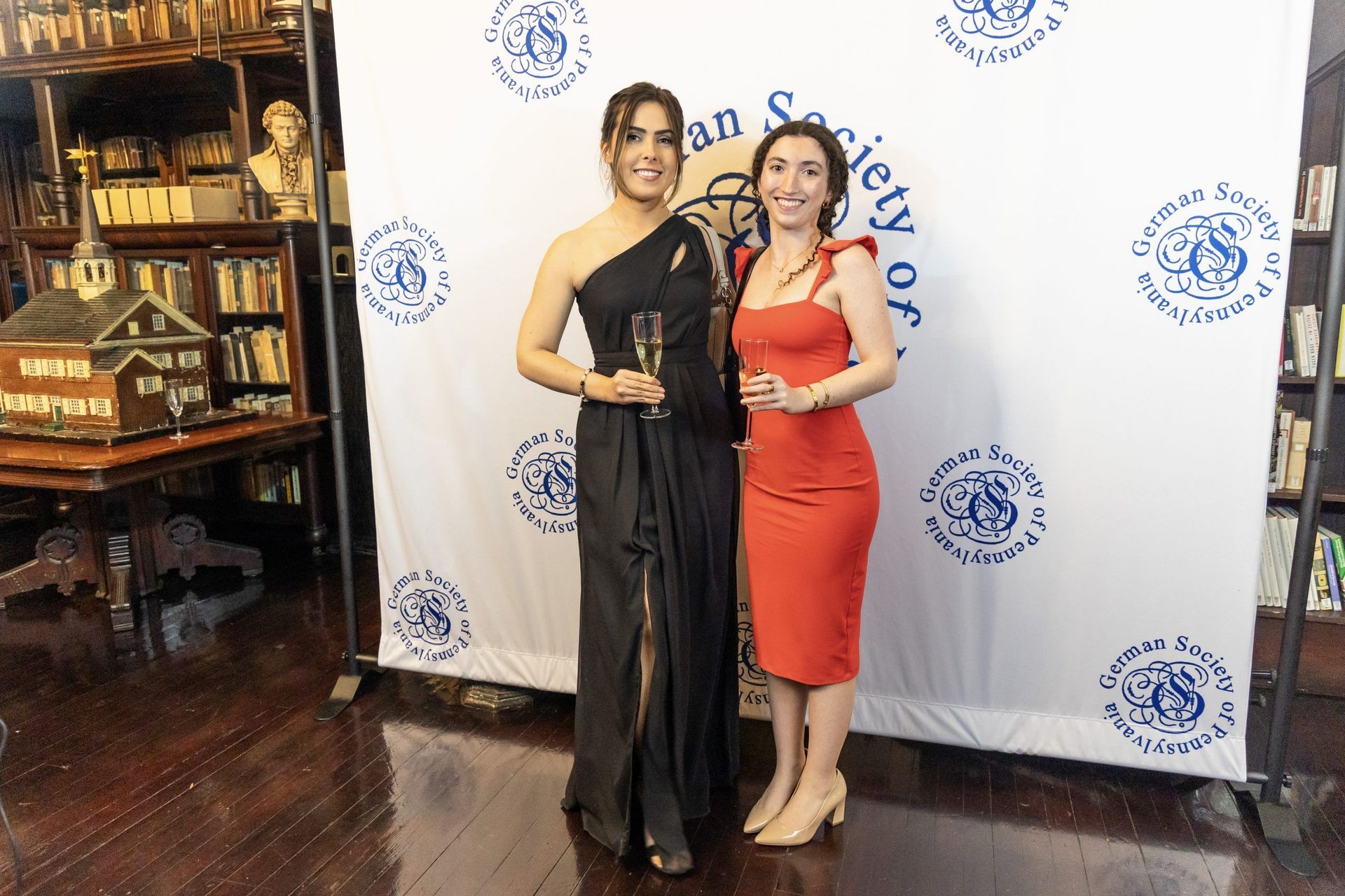 Two women posing at a formal event in front of a branded backdrop, one in a black gown and one in a red dress