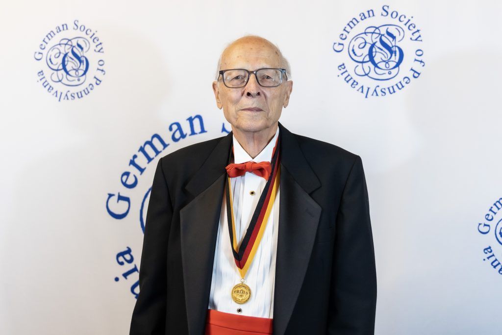 Man in tuxedo with medal standing before German Society backdrop