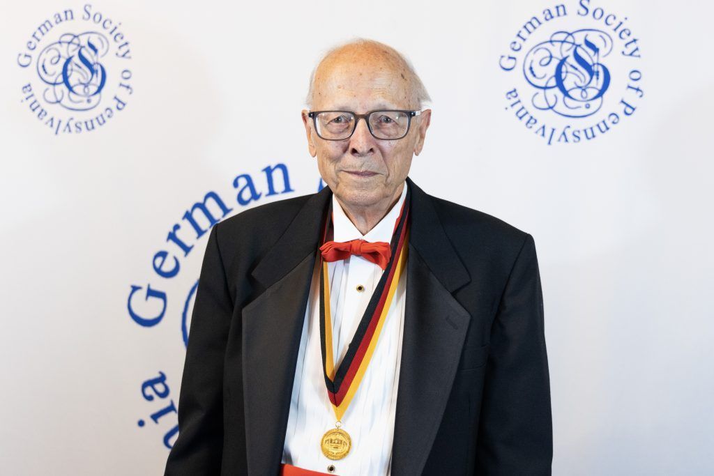 Elderly man in tuxedo wearing a medal, standing before a German Society backdrop