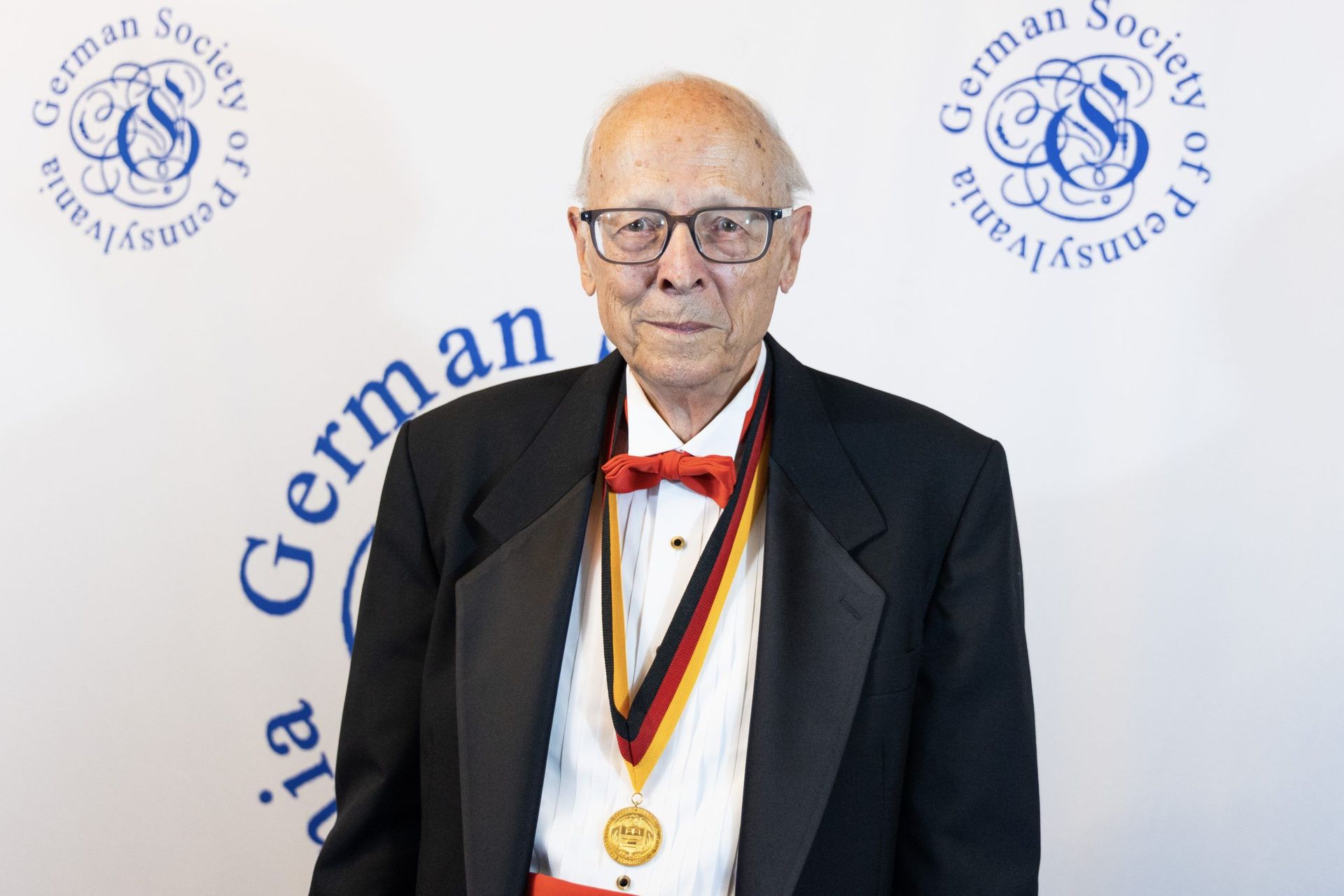 Elderly man in tuxedo with medals and bow tie standing before a German Society backdrop
